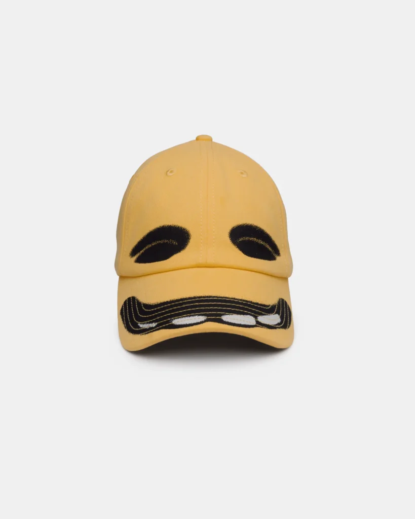 Pale yellow six-panel baseball cap embroidered with a stylized face graphic. Thick black curved eyebrows appear on the crown while a black-and-white moustache spans the front of the curved brim, creating a playful facial expression