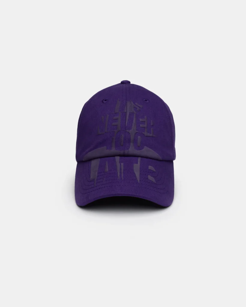 Deep purple baseball cap with tonal black oversized typography extending across the crown and brim. The layered graphic creates a bold, textured monochrome look while maintaining a minimalist color palette and curved visor