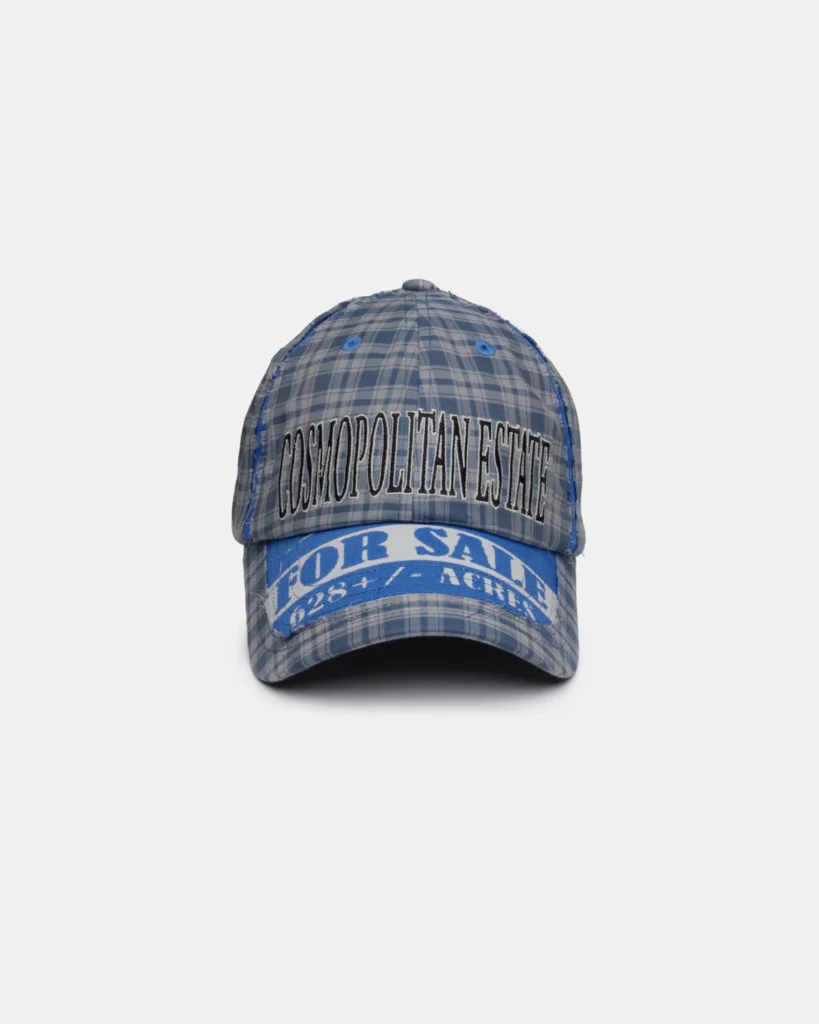 Blue and gray plaid baseball cap with distressed edges and embroidered "COSMOPOLITAN ESTATE" lettering across the front. A bold blue patch wraps around the brim with additional white typography, giving the cap a vintage-inspired aesthetic