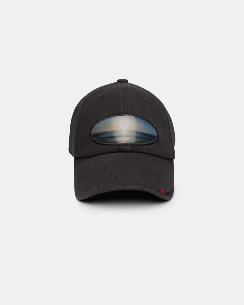 Charcoal gray baseball cap featuring an oval patch on the front displaying a serene ocean horizon at sunrise or sunset. A small red heart accent and subtle embroidered branding decorate the edge of the curved brim