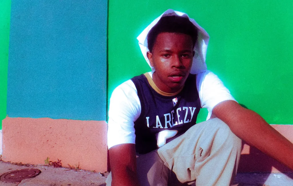 La Reezy leans against a vibrant wall in a striking portrait, wearing a 'LAREEZY' basketball jersey and hoodie, capturing the young New Orleans rapper's confident and introspective energy ahead of 'Family Bizzy