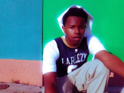 La Reezy leans against a vibrant wall in a striking portrait, wearing a 'LAREEZY' basketball jersey and hoodie, capturing the young New Orleans rapper's confident and introspective energy ahead of 'Family Bizzy
