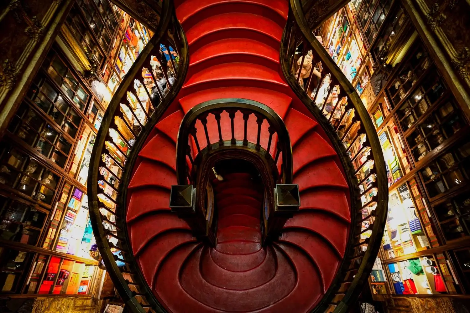 Dramatic view inside Livraria Lello in Porto, Portugal, showcasing its iconic sweeping crimson staircase surrounded by towering carved wooden bookshelves filled with books. Warm lighting highlights the ornate Gothic-inspired architecture, intricate railings, and symmetrical design, creating one of the world's most recognizable bookstore interior