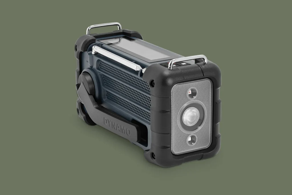 Rear three-quarter view of the portable emergency radio highlighting its integrated LED flashlight, fold-out dynamo hand crank, solar charging panel, and protective shock-resistant housing. The navy-blue body is wrapped with black rubber guards and metal carry handles, emphasizing its preparedness-focused design on a clean olive-green backdrop