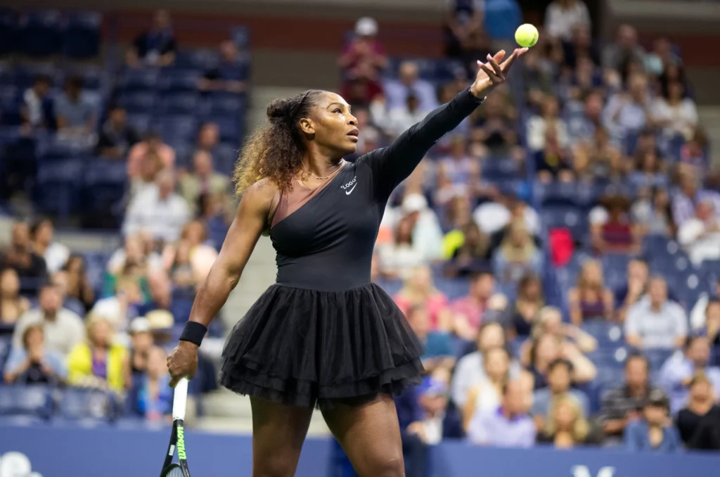 Tennis player tosses the ball high during a serve at a packed stadium, wearing a black one-shoulder dress with layered skirt detailing. The action shot captures focus, power, and elegance before the point begin