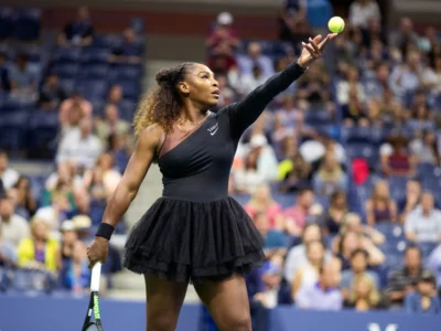 Tennis player tosses the ball high during a serve at a packed stadium, wearing a black one-shoulder dress with layered skirt detailing. The action shot captures focus, power, and elegance before the point begin