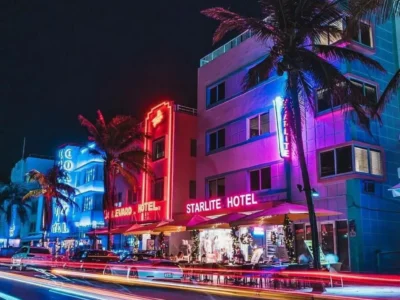 A vibrant nighttime streetscape captures the heart of Miami's Art Deco district, where iconic pastel hotels glow beneath brilliant neon lighting in shades of blue, pink, and red. Towering palm trees frame the illuminated facades, while long-exposure light trails from passing traffic streak across the foreground, creating a dynamic sense of movement. The retro architecture, electric signage, and tropical atmosphere evoke classic 1980s South Florida aesthetics, blending nostalgic glamour with the city's unmistakable nightlife energy