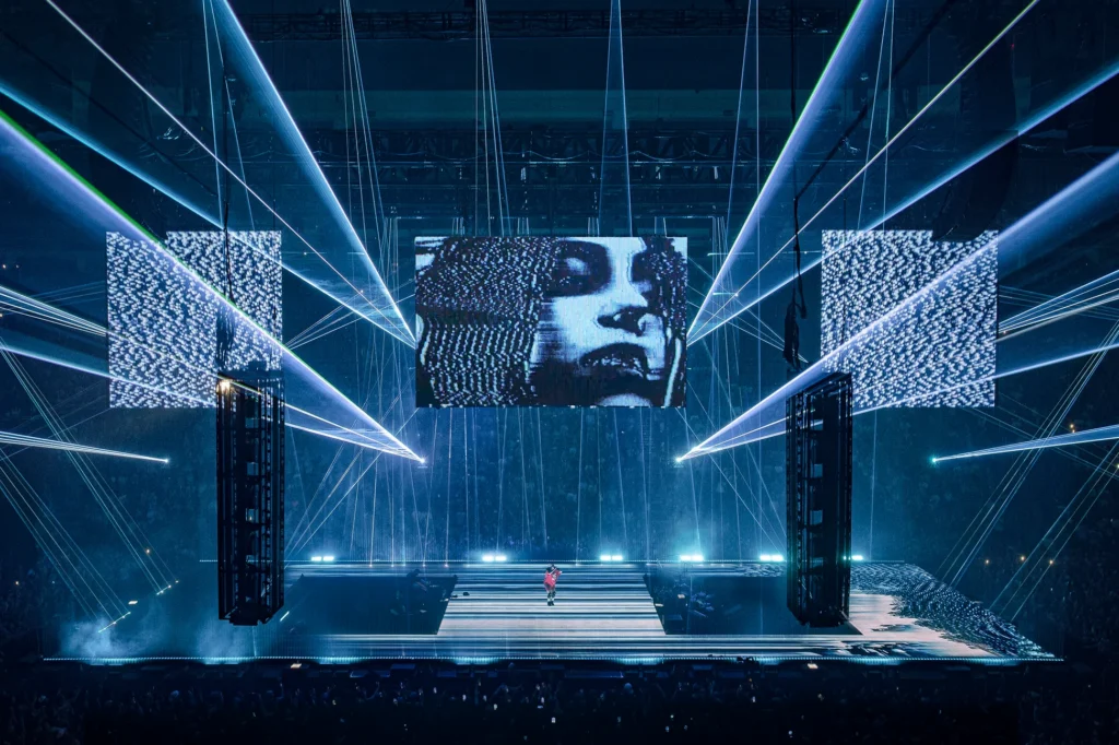 Wide view of a large-scale concert stage featuring intersecting blue laser beams, towering suspended LED screens, and a central glitch-art portrait displayed above the performer. The futuristic production combines immersive lighting, digital visuals, and dramatic stage architecture inside a packed indoor arena