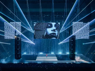 Wide view of a large-scale concert stage featuring intersecting blue laser beams, towering suspended LED screens, and a central glitch-art portrait displayed above the performer. The futuristic production combines immersive lighting, digital visuals, and dramatic stage architecture inside a packed indoor arena