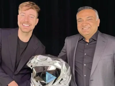 Two men stand side by side in front of a black backdrop, smiling while holding a large silver YouTube-style creator award. Both wear dark blazers over black shirts, creating a sleek, formal look. The reflective trophy, featuring a blue play-button centerpiece, dominates the foreground and symbolizes a major milestone in digital media and online content creation