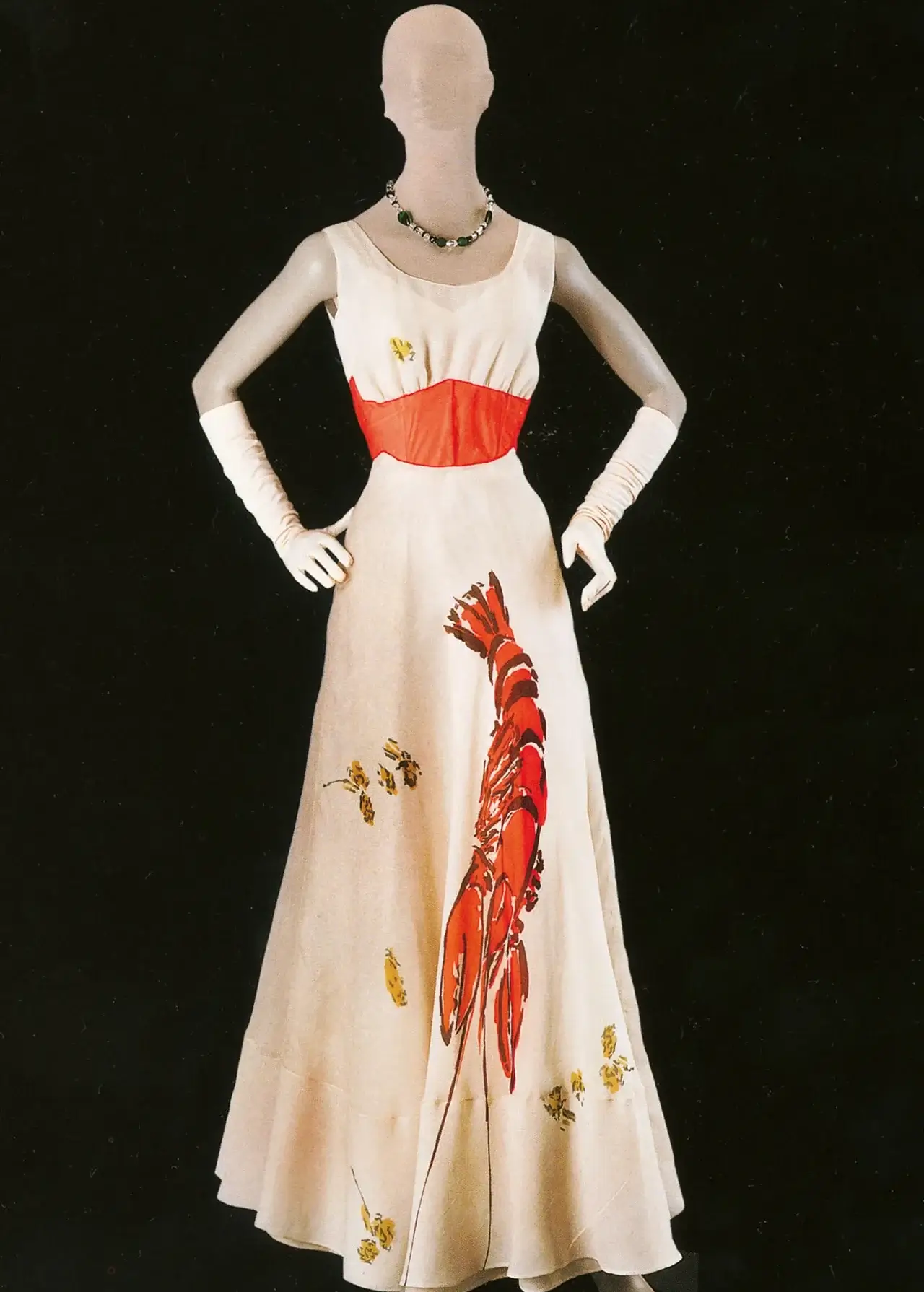 Archival Schiaparelli evening gown displayed on a mannequin, featuring a striking red lobster graphic across the skirt and a coral-tincture waist panel against an ivory sil