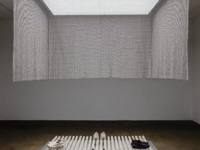 Wide view of a minimalist exhibition space featuring a suspended metallic mesh canopy above a white platform filled with rows of white dandelion seed heads mounted on slender rods. Several On footwear models are displayed among the installation, creating a sculptural presentation beneath soft overhead light