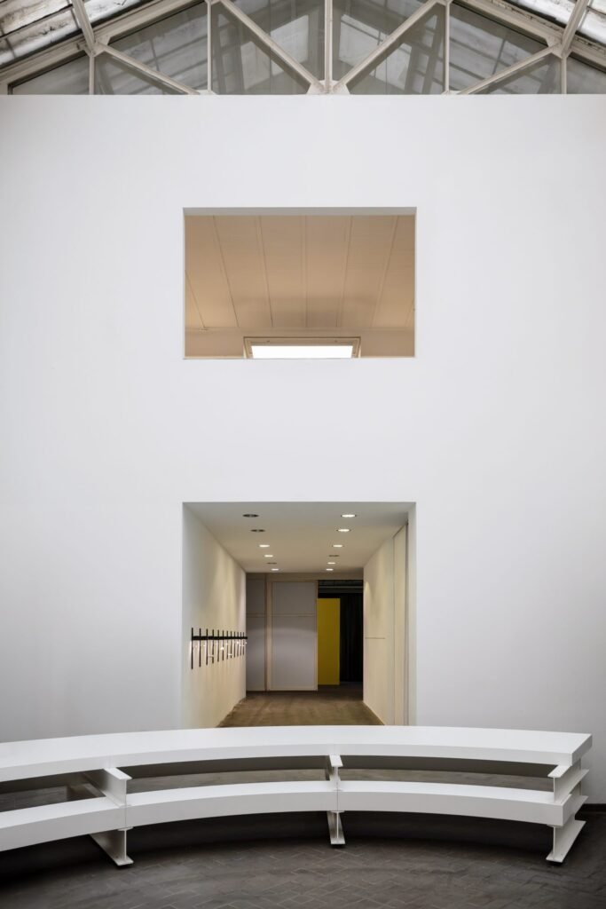 Minimalist gallery interior with a large white wall, a rectangular viewing opening, and curved white tiered seating arranged in the foreground. Recessed lighting and clean architectural lines emphasize the spacious, contemporary showroom environment
