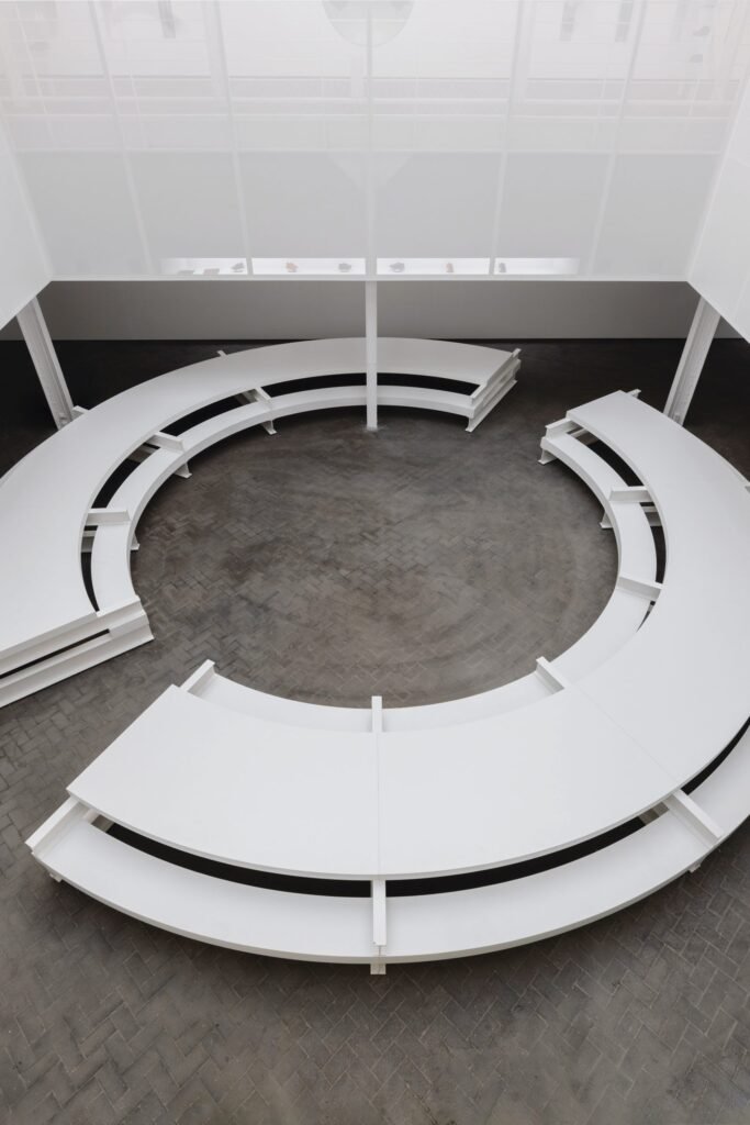 Overhead perspective of the circular white seating installation surrounding an open central floor space. The translucent suspended partition and skylit industrial architecture frame the minimalist exhibition layout