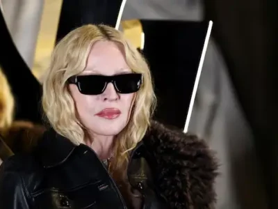 Madonna wearing a black leather jacket and dark sunglasses, posing confidently with softly waved blonde hair against a dramatic black-and-gold backdrop at a fashion and entertainment event