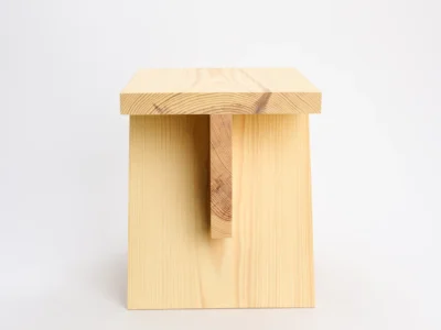 Front-facing studio photograph of the pinewood stool against a light gray backdrop. The design showcases a solid rectangular seat supported by two vertical side panels and a central structural brace beneath the top. Visible wood grain and natural knots emphasize the material’s organic character and craftsmanship