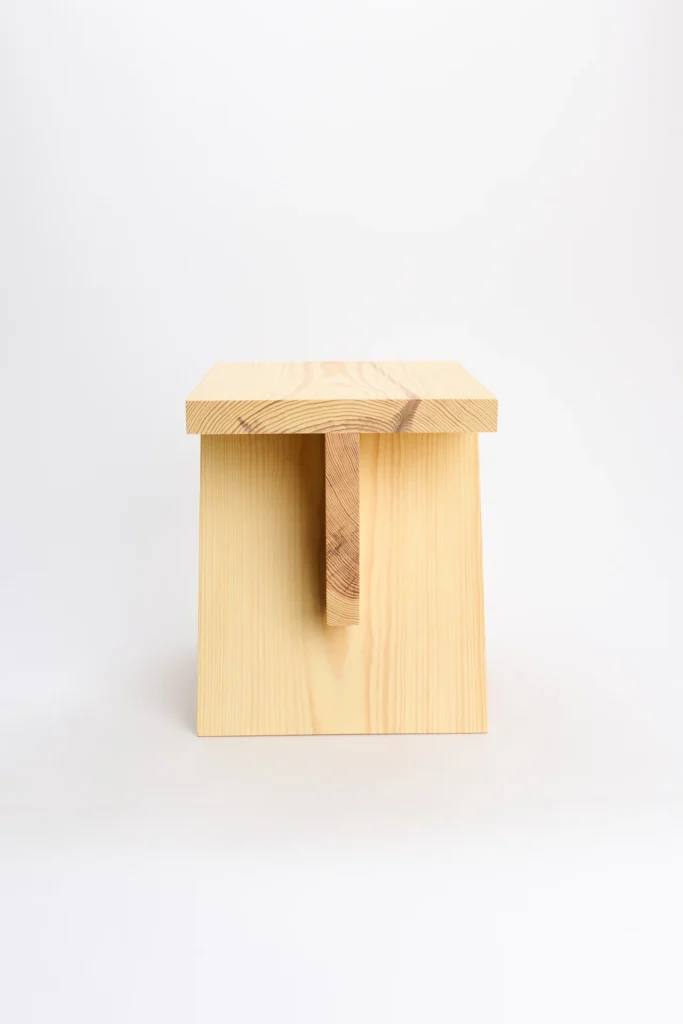 Front-facing studio photograph of the pinewood stool against a light gray backdrop. The design showcases a solid rectangular seat supported by two vertical side panels and a central structural brace beneath the top. Visible wood grain and natural knots emphasize the material’s organic character and craftsmanship