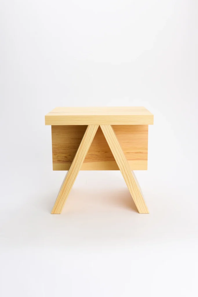 Rear view of the stool highlights the distinctive A-frame leg structure positioned beneath the rectangular seat. A suspended wooden panel spans between the legs, creating a balanced composition of straight and angled forms. Natural grain patterns run consistently across the solid pine surfaces, emphasizing the handcrafted construction