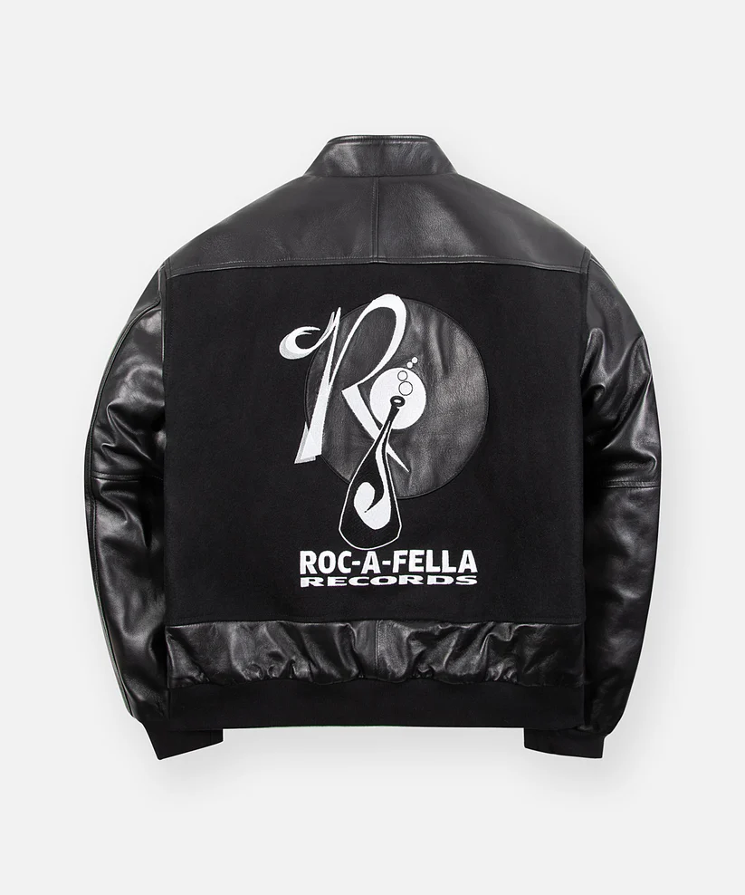 Rear view of the black varsity jacket highlighting an oversized embroidered Roc-A-Fella Records logo across the back panel. The black-and-white graphic dominates the wool body, framed by leather shoulders and sleeves, reinforcing the collection's tribute to Jay-Z's influential debut era