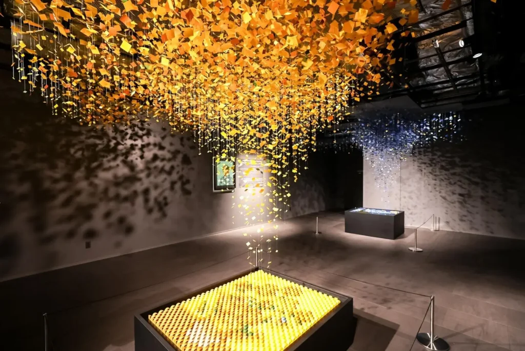 Suspended golden paper-like elements cascade from the ceiling in a contemporary art installation, casting intricate sil throughout a dark gallery room