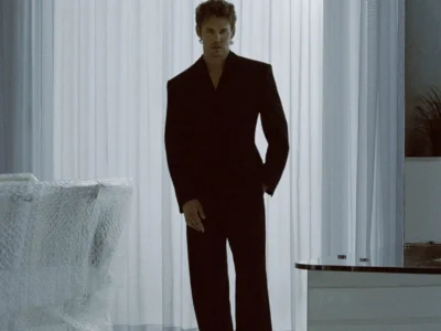 Model in a sharply tailored black suit stands in a minimalist white room, framed by sheer curtains and reflective flooring, creating a cinematic Saint Laurent atmosphere