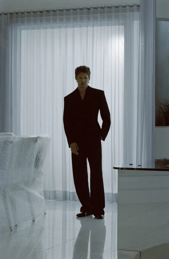Model in a sharply tailored black suit stands in a minimalist white room, framed by sheer curtains and reflective flooring, creating a cinematic Saint Laurent atmosphere