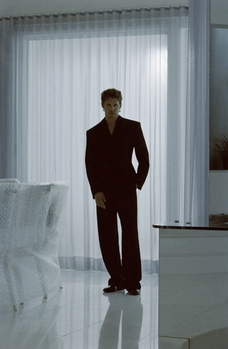 Model in a sharply tailored black suit stands in a minimalist white room, framed by sheer curtains and reflective flooring, creating a cinematic Saint Laurent atmosphere