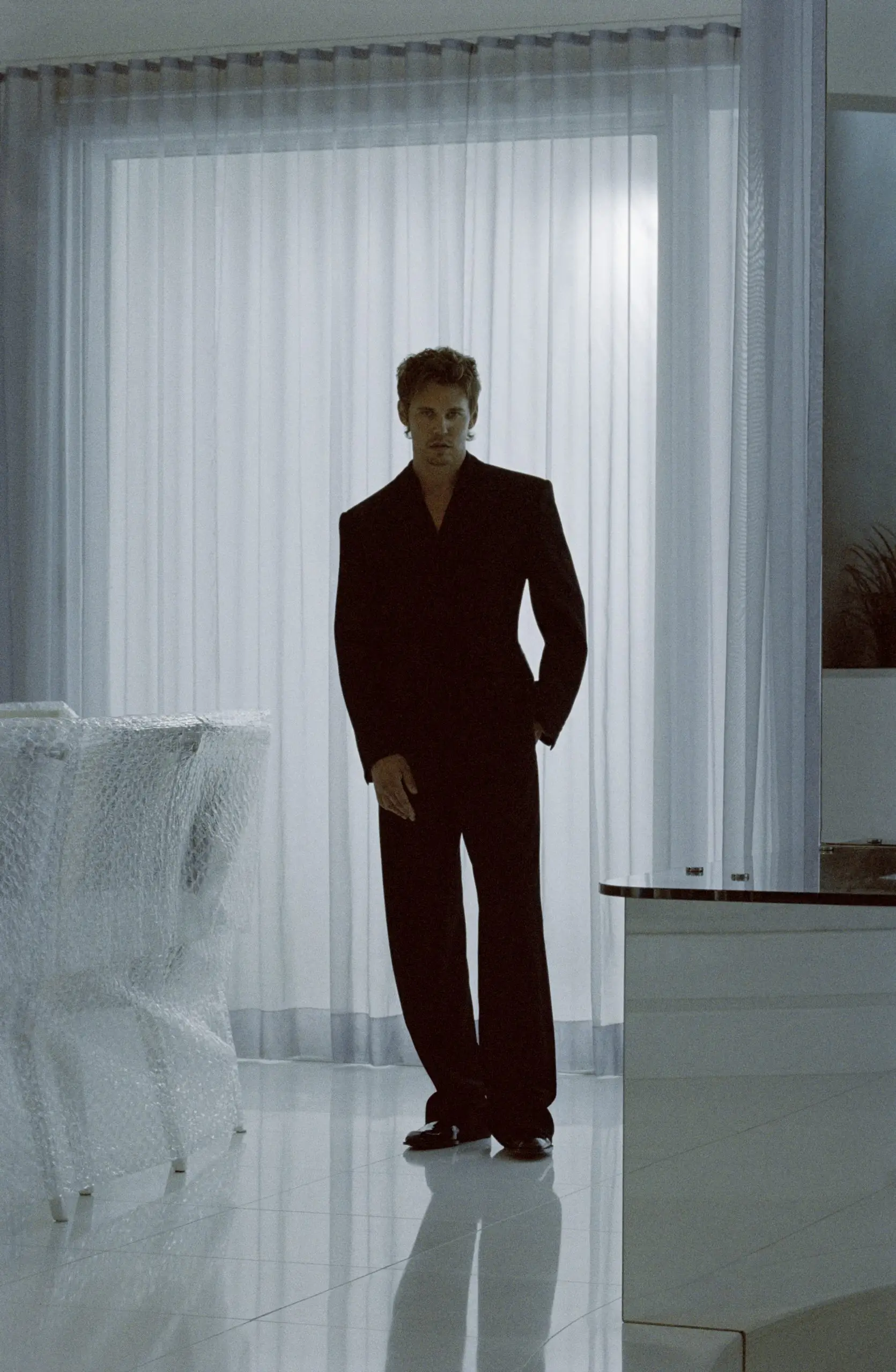 Model in a sharply tailored black suit stands in a minimalist white room, framed by sheer curtains and reflective flooring, creating a cinematic Saint Laurent atmosphere