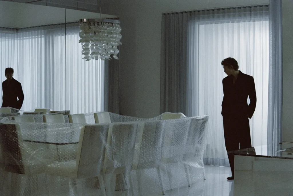 Model in an oversized black suit poses beside a long dining table wrapped in bubble wrap, surrounded by mirrors and soft natural light