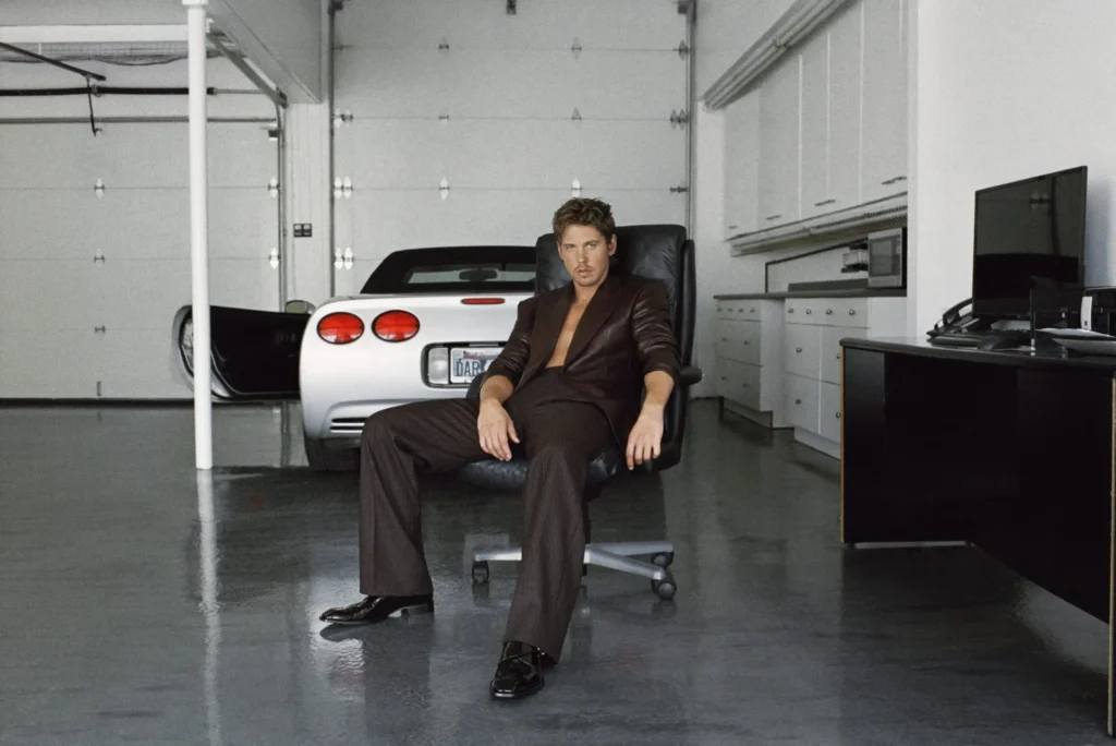 Model seated in a black office chair inside a pristine garage, wearing an open black suit with a white sports car parked behind him