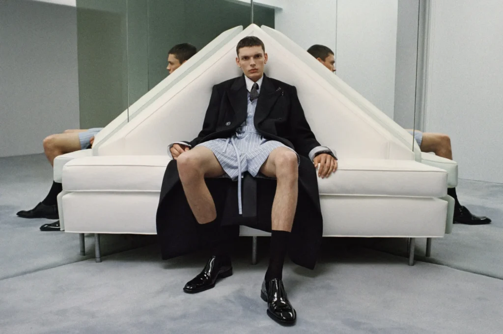 Model sits on a geometric white sofa wearing an oversized black coat, striped shorts, tie, and polished loafers in a mirrored room