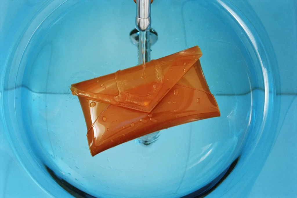 Orange patent leather envelope clutch suspended beneath running water inside a circular blue basin, emphasizing texture and shine
