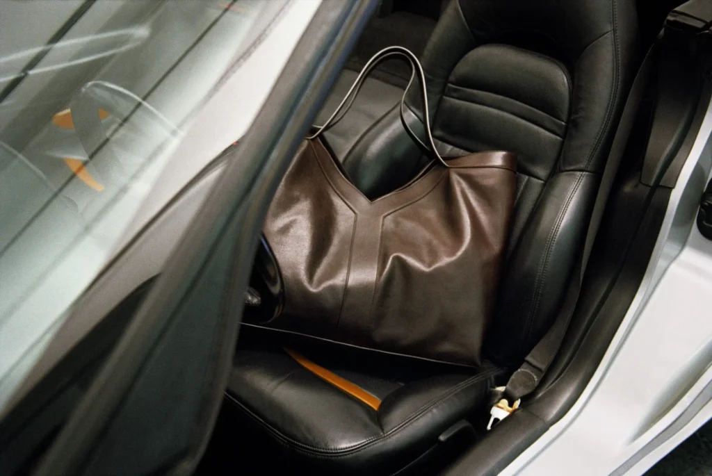 Large brown leather tote bag rests on the passenger seat of a silver sports car, photographed in a luxury garage setting