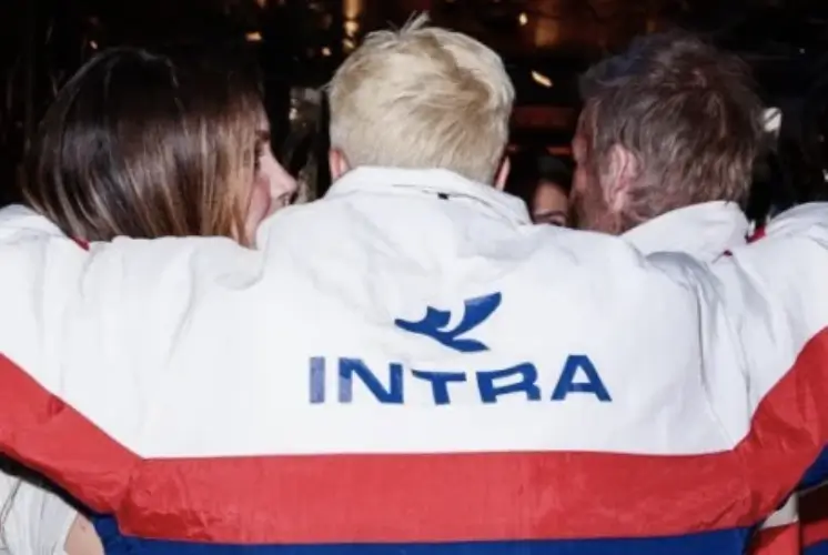Three people stand closely together with their arms around each other at a nighttime gathering. The central figure wears a white, red, and blue jacket featuring a large "INTRA" logo across the back, while warm ambient lights and a crowded social setting create a candid atmosphere of friendship, community, and connection