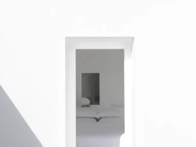 Minimalist whitewashed Santorini residence framed by a clean geometric doorway, revealing a serene bedroom beyond with soft natural light and a palm tree rising above the courtyard wall