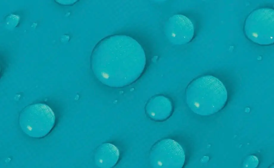 Extreme close-up of a bright blue waterproof fabric surface covered with rounded water droplets. The beads of water sit on top of the material rather than soaking in, highlighting the fabric’s water-repellent coating and outdoor-performance functional