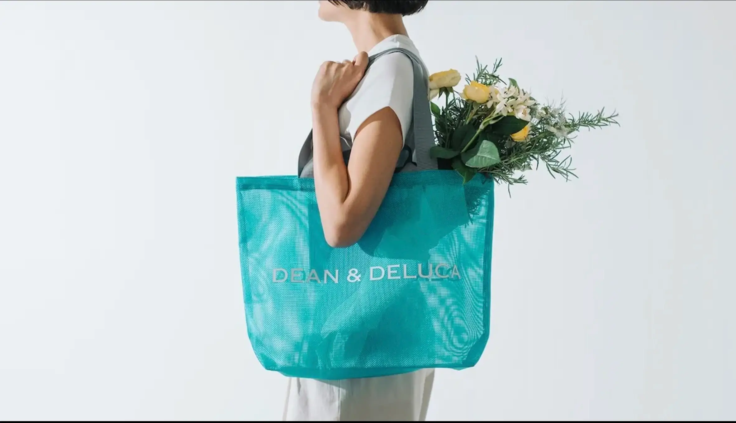 A person carrying a large turquoise DEAN & DELUCA mesh tote filled with fresh flowers. The translucent bag features gray handles and white logo branding, photographed in a bright, minimalist interior with soft natural light