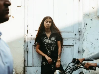 A portrait framed by passing pedestrians and motorcycles, showing a model posed in front of distressed white wooden doors. Her dark embroidered outfit contrasts with the faded architecture, blending contemporary fashion with an authentic street environment