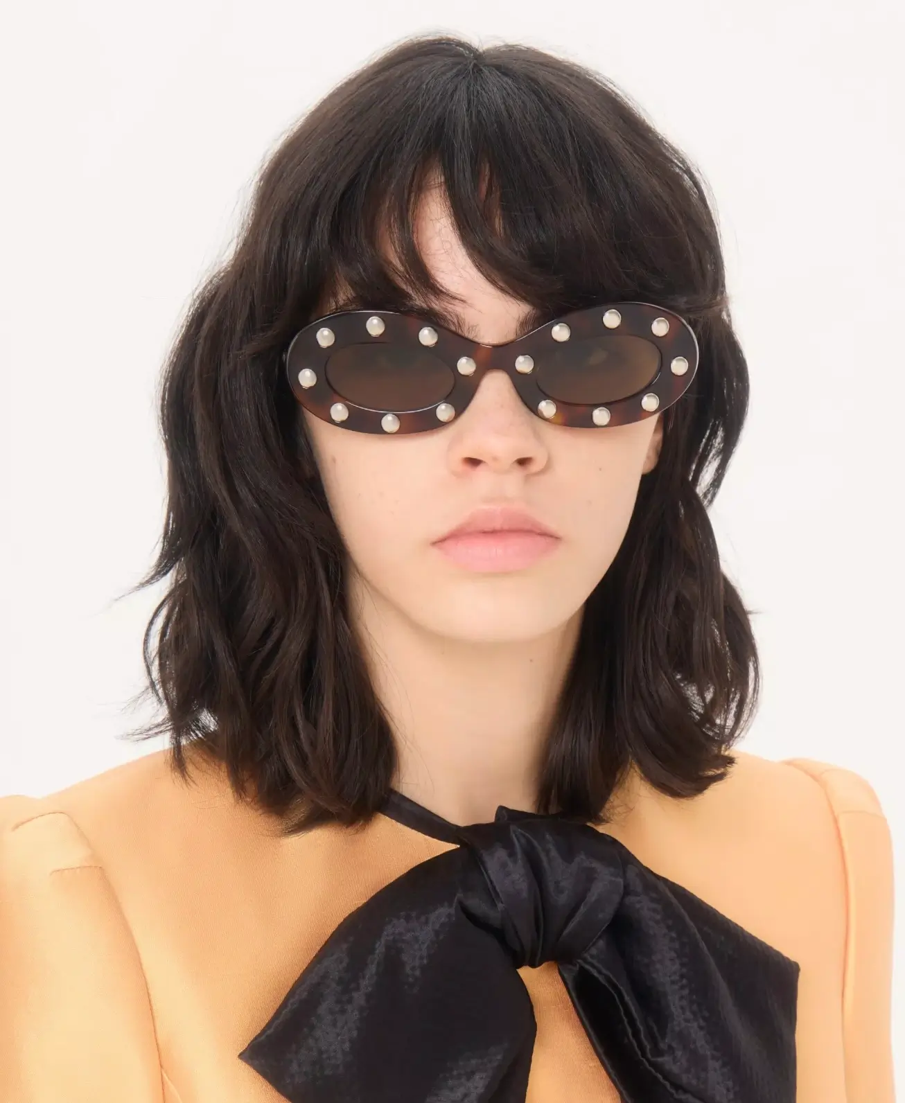 Model with shoulder-length dark hair wearing Valentino tortoiseshell oval sunglasses accented with silver studs, paired with a peach-toned tailored jacket and oversized black bow detail
