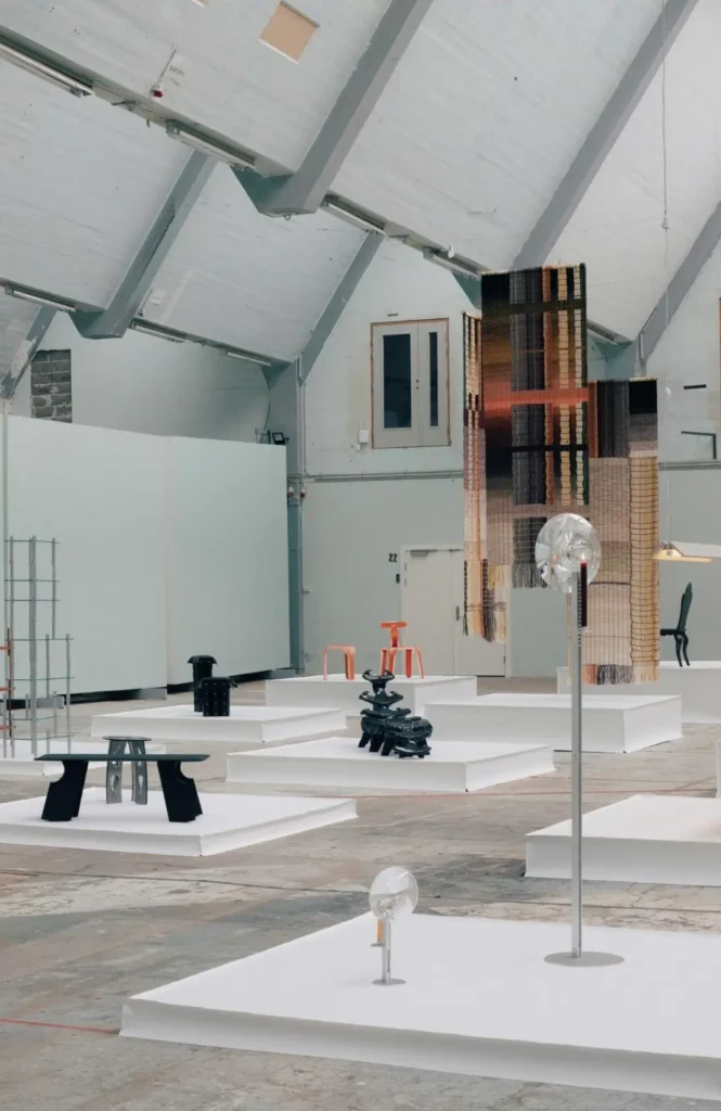 Wide view of a contemporary design exhibition featuring furniture, lighting, and textile installations displayed on white plinths inside a bright warehouse-style hall with exposed beams and high ceiling