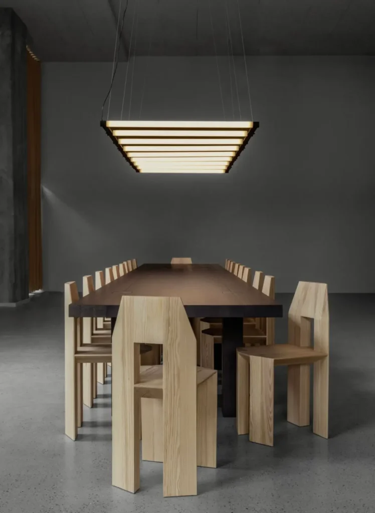Minimalist dining installation with a long dark wood table surrounded by angular pale-wood chairs beneath a layered rectangular pendant light, creating a calm architectural atmosphere