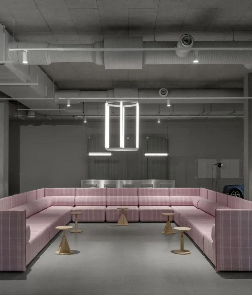 Large U-shaped sectional sofa upholstered in pink plaid fabric, arranged in an industrial showroom beneath suspended geometric lighting and surrounded by small wooden side table