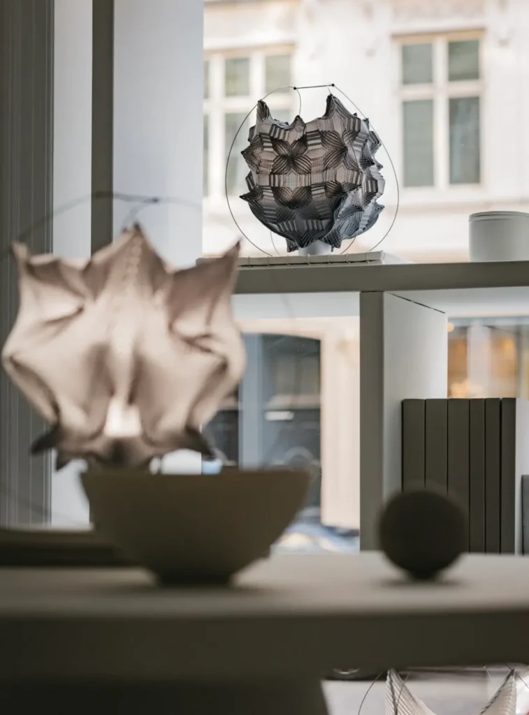 Delicate textile sculptures displayed on shelving near large windows, showcasing intricate folded forms and translucent woven materials against a softly lit urban backdrop