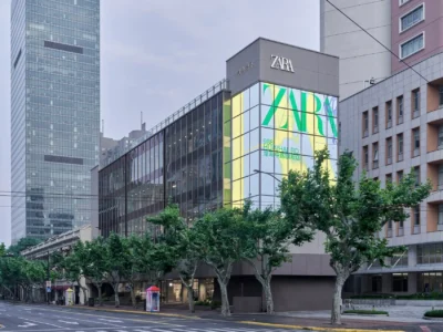 A multi-story flagship retail building with a glass façade and prominent branding. Floor-to-ceiling windows reveal expansive interior spaces, while tree-lined streets and surrounding high-rises situate the store within a vibrant urban environment