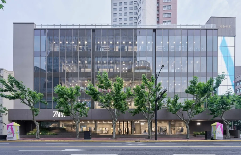 A contemporary flagship storefront featuring a transparent glass façade that showcases multiple levels of retail space. Bright interior lighting, modern architecture, and a landscaped streetscape create a strong visual presence within the city