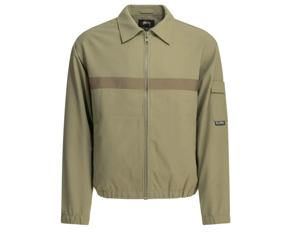 Front view of a khaki Stüssy jacket with a full-zip closure, pointed collar, elasticized hem and cuffs, and a contrasting horizontal webbing stripe across the chest