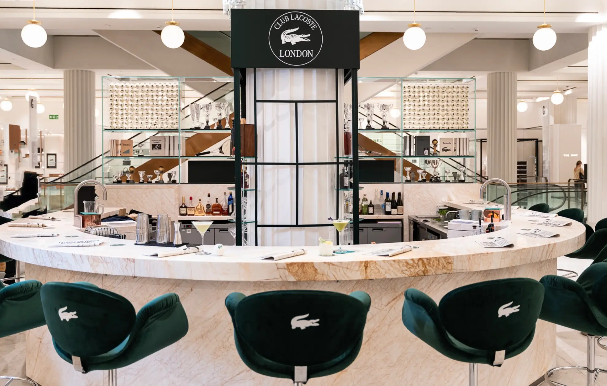 Symmetrical view of the circular Club Lacoste bar with marble finishes, green velvet seating, curated cocktails, and trophy displays set within a luxury retail destination