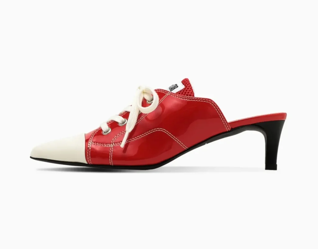 Clean lateral view of the patent red mule showcasing classic shoe-inspired eyelets, cream laces, white stitching details, and a sharply pointed toe balanced on a black heel