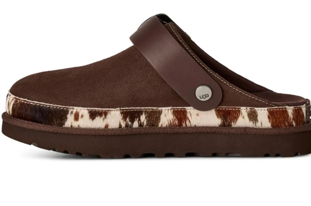 Side profile of a UGG suede slingback clog in dark brown with a smooth leather heel strap and silver hardware. A cow-print calf hair trim wraps around the midsole, contrasting with the suede upper and chunky outsole