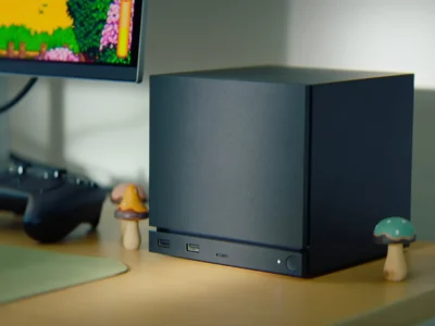 Compact black cube-shaped Steam Machine gaming PC positioned on a wooden desk beside a monitor displaying colorful pixel-art gameplay. The minimalist matte-black chassis features front-facing USB ports and a power button, highlighting Valve’s modern living-room gaming hardware design that blends console simplicity with desktop PC show